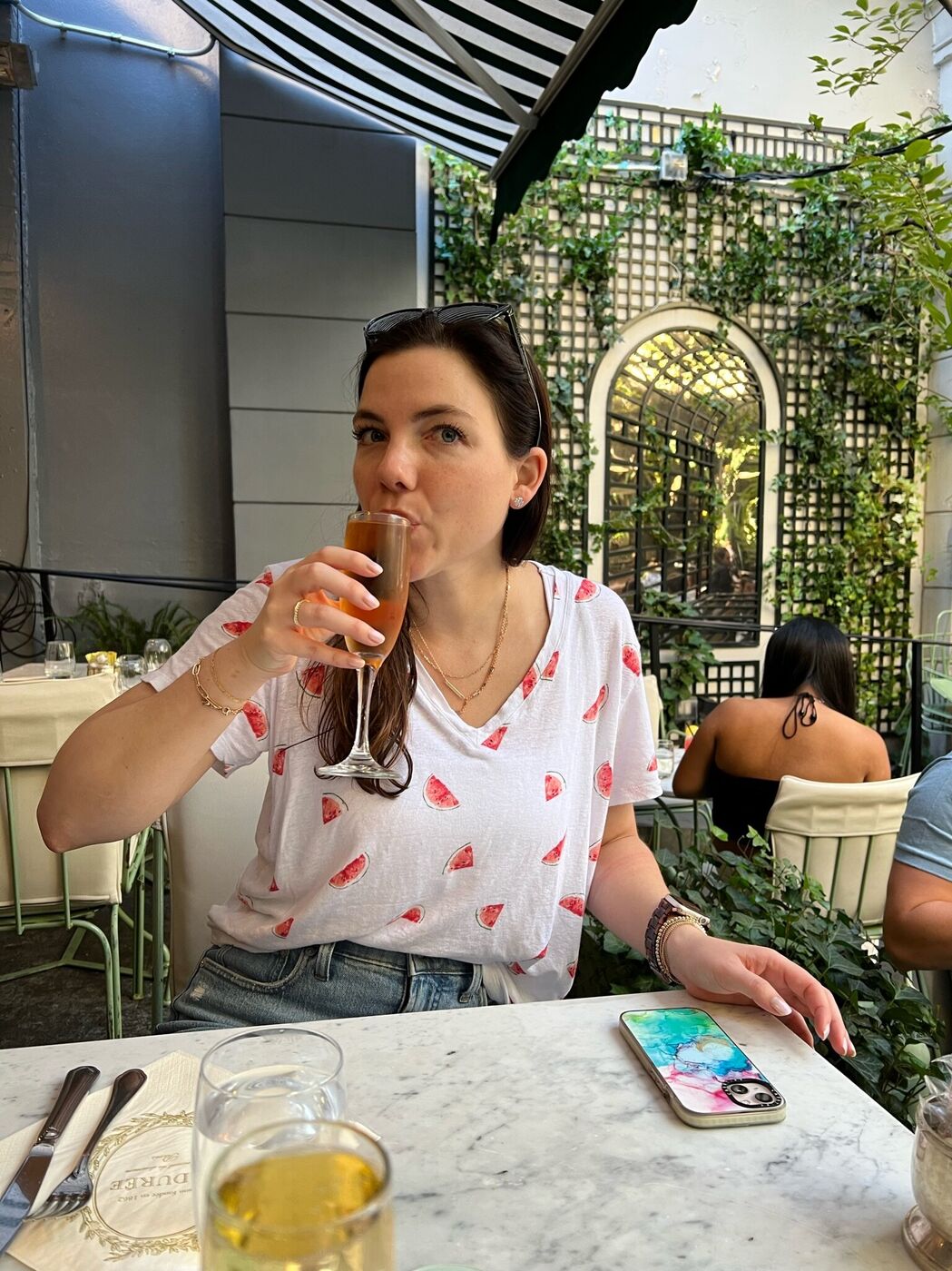 Elainea sipping a mimosa on a café patio in a watermelon-print tee